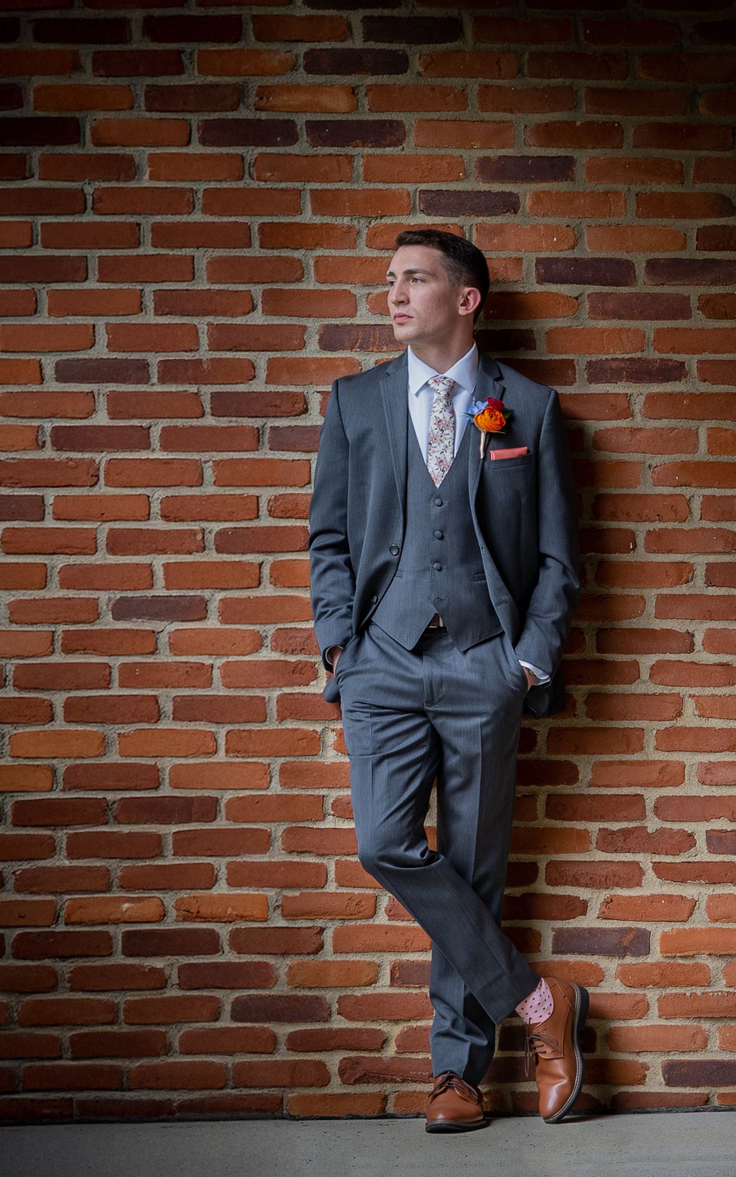Nationwide Hotel & Conference Center Groom in gray suit poses casually against rustic brick wall with orange boutonniere.