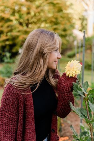 Person in burgundy knit sweater leans to smell yellow dahlia flower in autumn garden setting.