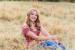 A person in a burgundy top and jeans sits casually in a golden autumn field during sunset.