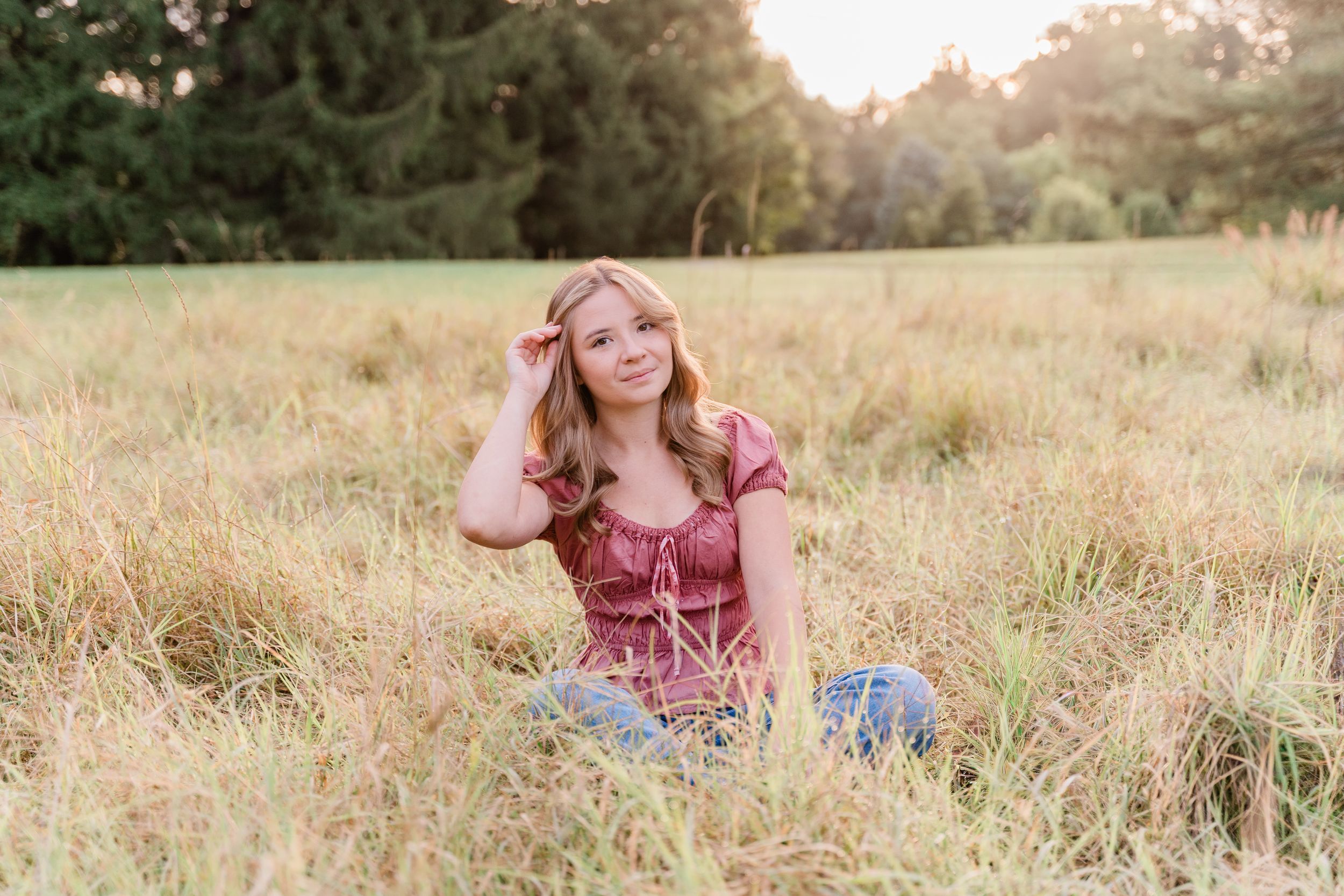A figure in a burgundy dress sits gracefully in a sunlit meadow during golden hour.