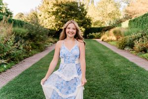 A person in a blue floral sundress twirls joyfully in a sunlit garden path during summer.