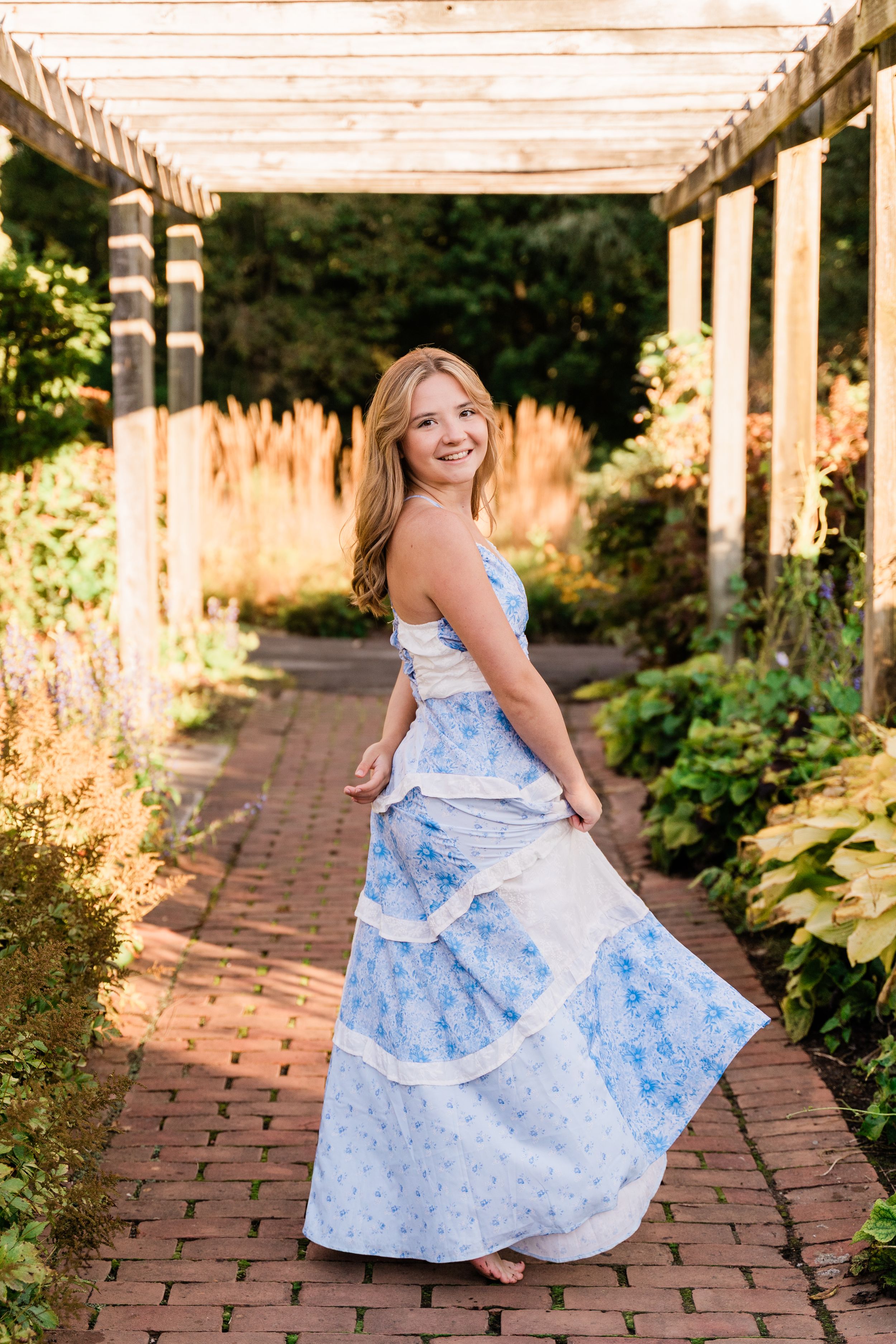 A child in a blue floral dress twirls joyfully along a brick pathway under a garden pergola in golden sunlight.