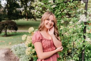 Woman in coral dress poses near blooming white flowers in a garden setting.