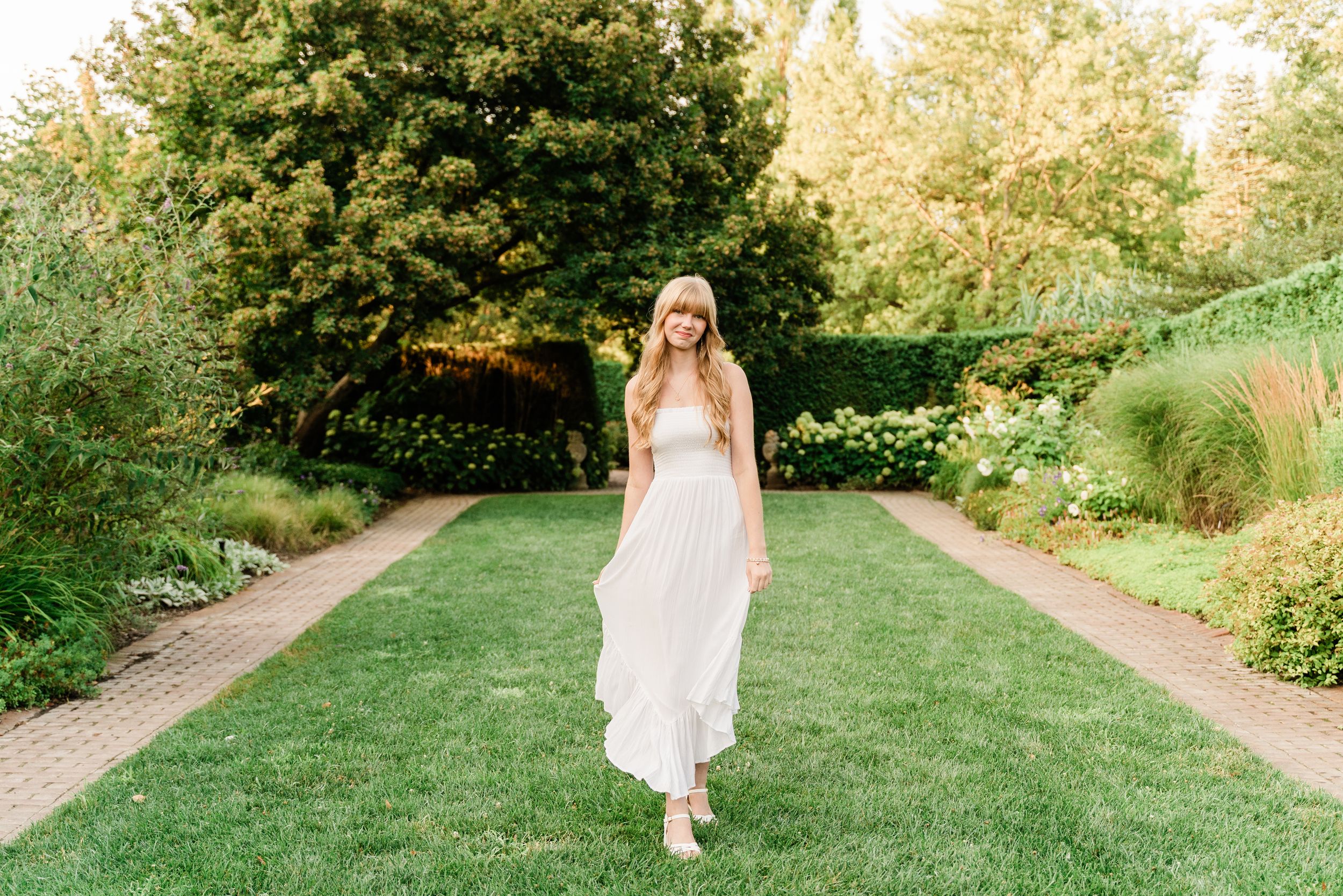 A bride in a flowing white dress walks along a garden path surrounded by lush greenery on a sunny day.