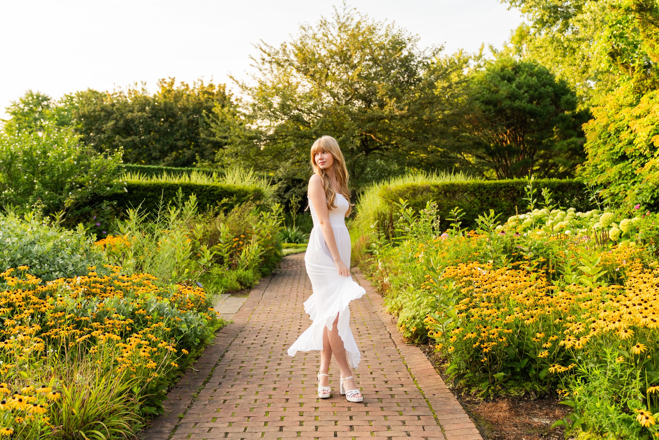 A person in a white dress twirls on a brick path surrounded by vibrant summer garden flowers and greenery.