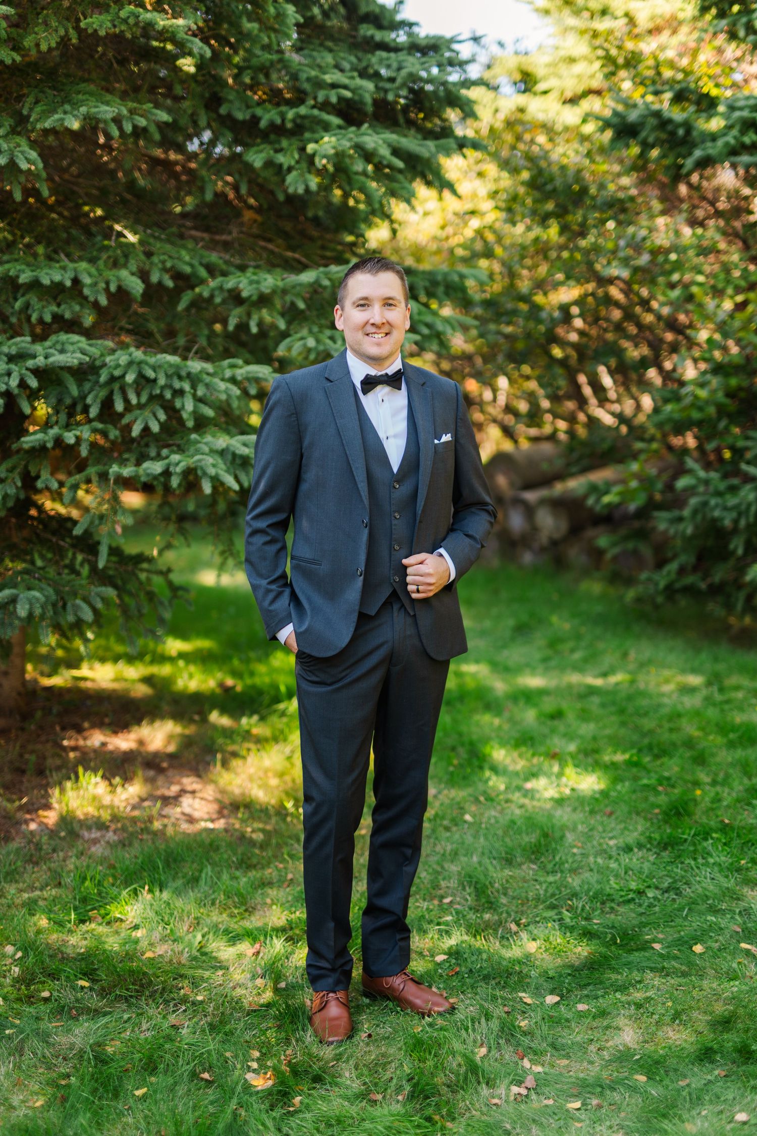 A groom in a classic black tuxedo and bow tie poses outdoors in a garden setting with lush green foliage.