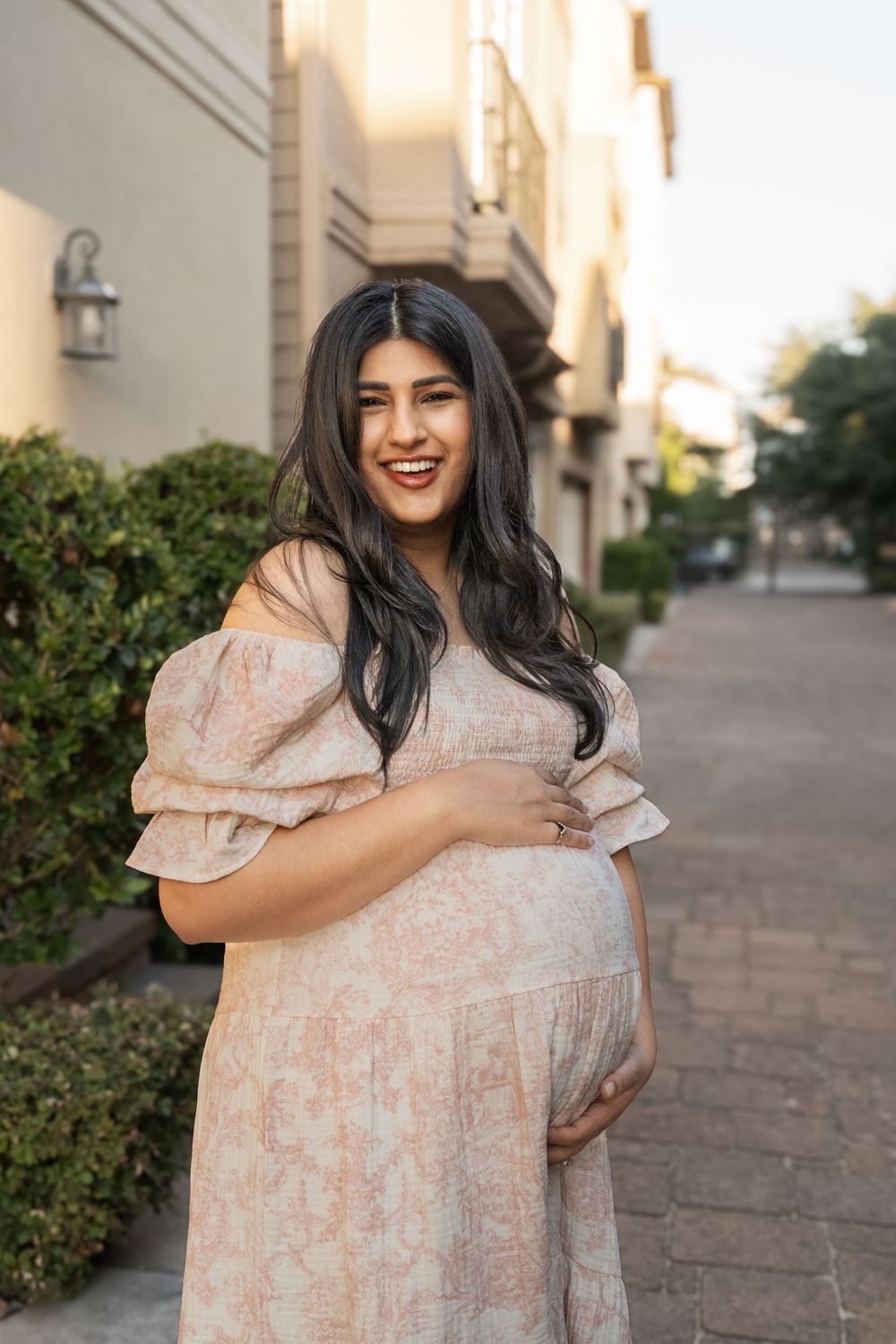 Expectant mother in peach dress smiling while standing on brick pathway.