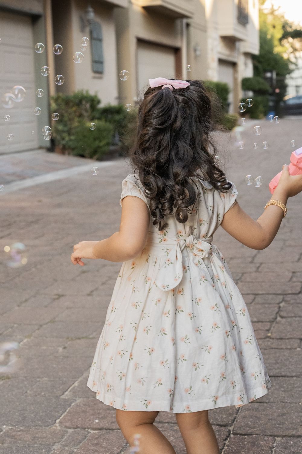 Young child in white floral dress chasing bubbles on a brick pathway in a playful sequence.