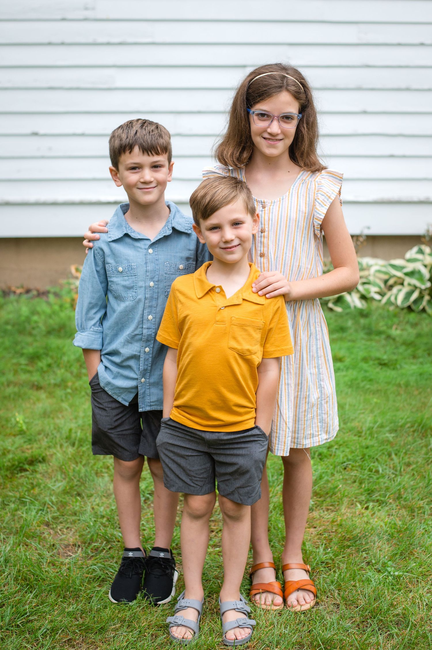 Three children stand together on grass in front of white siding wearing casual summer clothes and sandals.