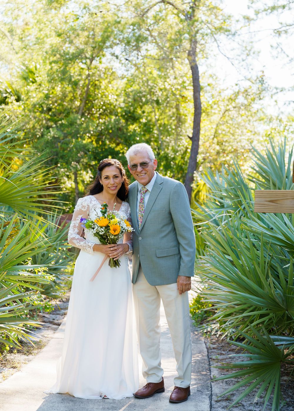 Wedding couple walks along garden path surrounded by tropical palm plants in sunlit outdoor setting.