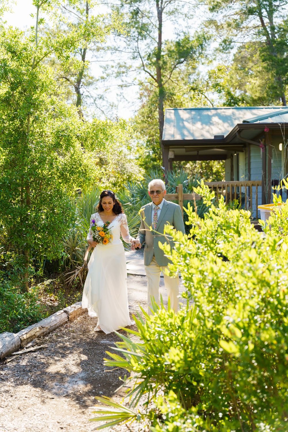 Couple walking along a garden path surrounded by lush greenery and sunlit foliage at an outdoor venue.