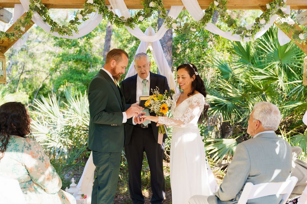 A wedding ceremony takes place in a lush outdoor garden setting with tropical plants and white decorations.