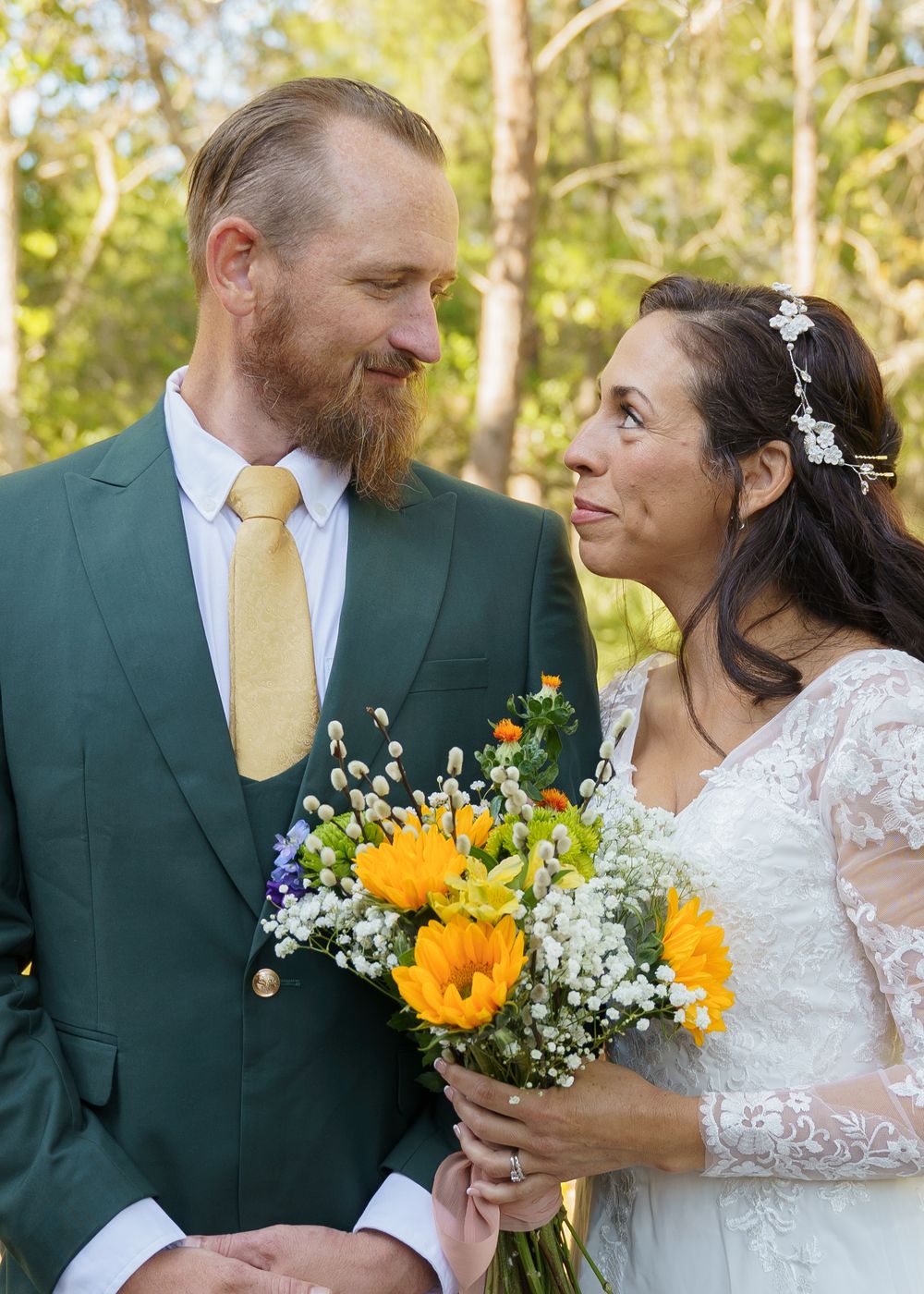 A couple in wedding attire share a tender moment outdoors, with the bride holding a vibrant sunflower and baby's breath bouquet.