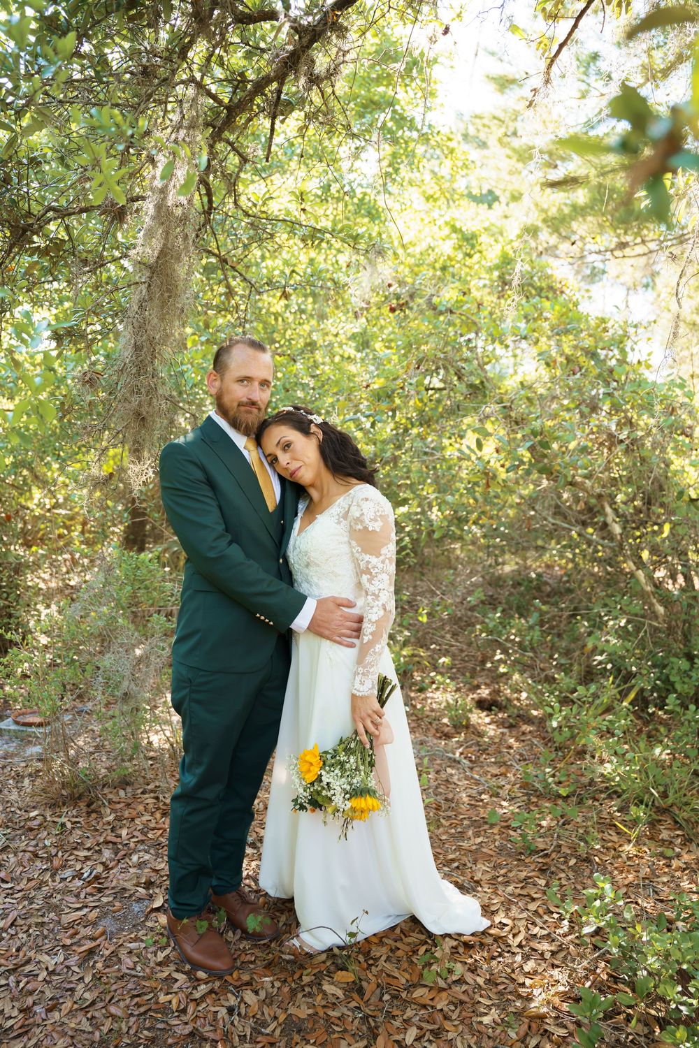 A romantic wedding couple embraces in a sunlit wooded area with natural greenery surrounding them.