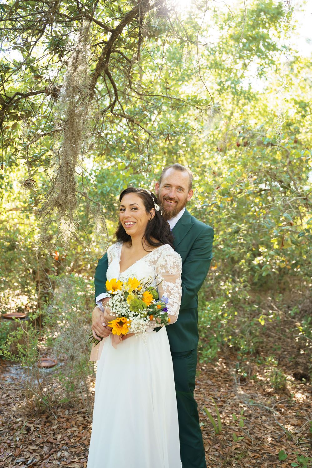 A romantic outdoor wedding photoshoot showing a couple embracing among sunlit trees with sunflowers and white lace details.
