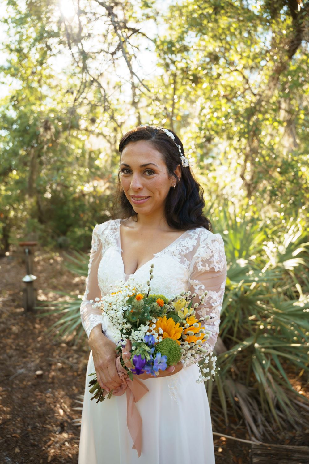 Bride in white lace dress holds vibrant sunflower and wildflower bouquet in garden setting.