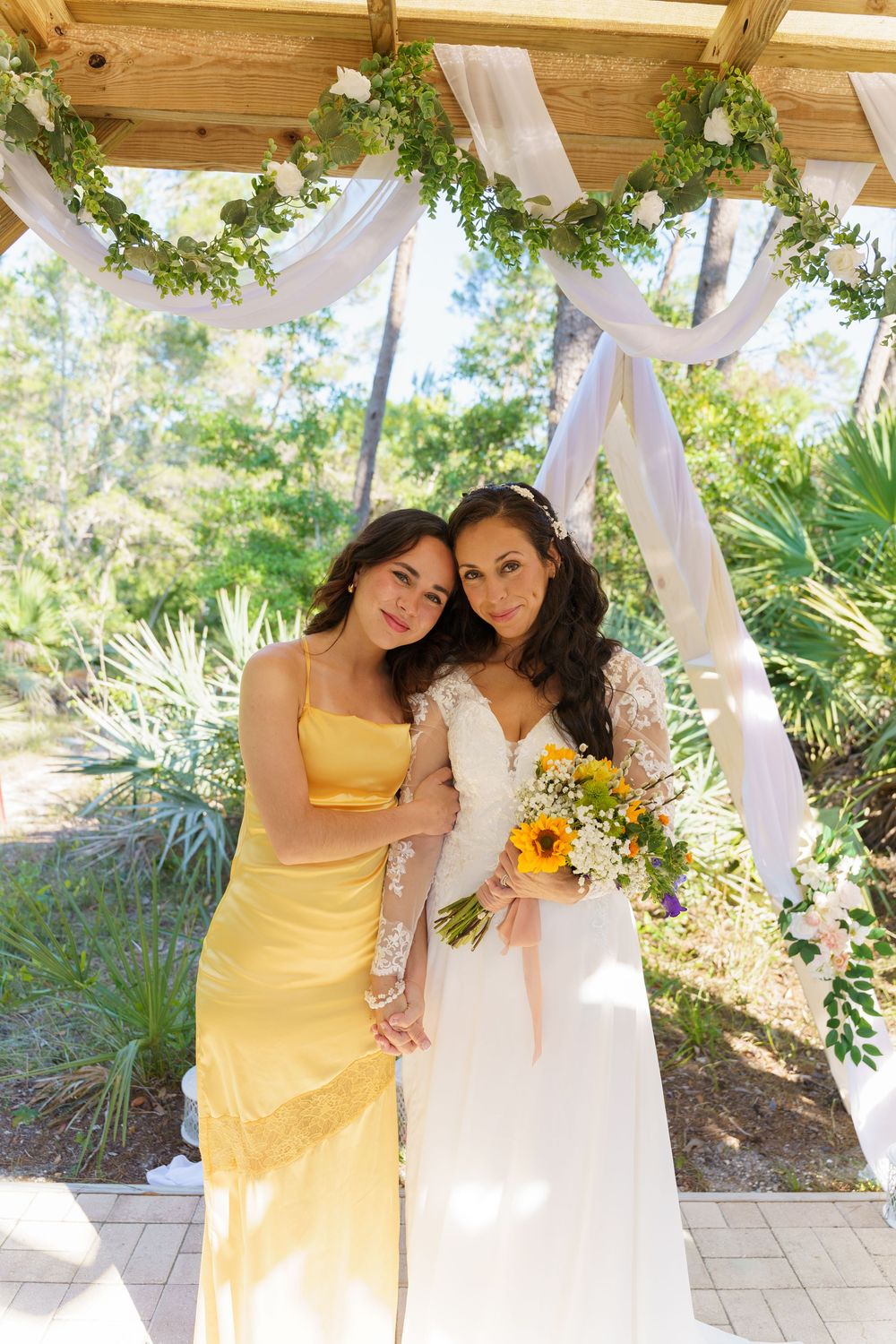 Wedding party poses under white draped pergola decorated with greenery and sunflowers.