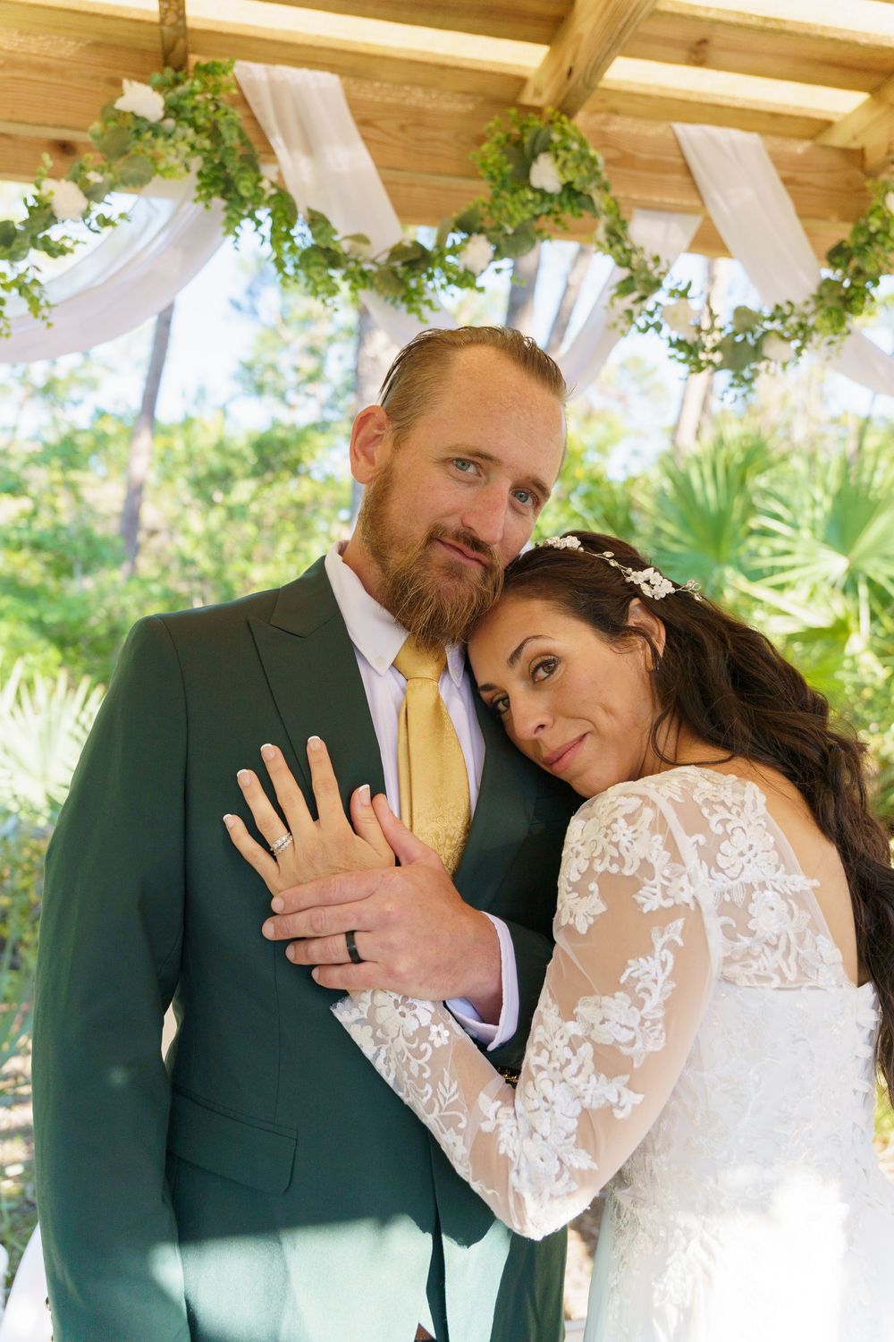 A bride in a lace sleeve wedding dress and groom in dark suit pose under a floral decorated wooden pergola.