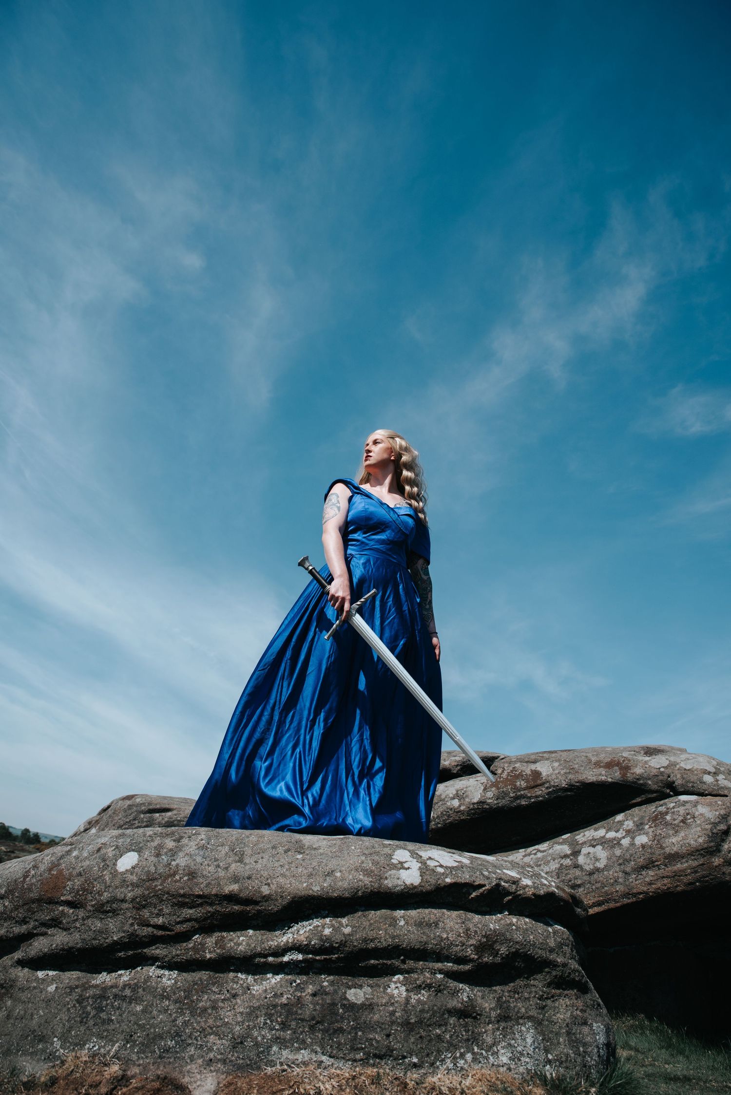 Figure in flowing blue dress posed dramatically on rocky outcrop against bright sky.