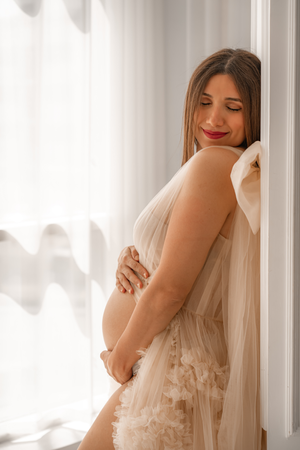 Dreamy boudoir maternity photos with soft window light and sheer white curtains.