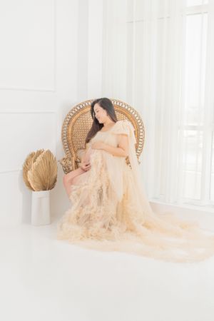 Maternity portrait in peachy lace dress on wicker chair with soft lighting.