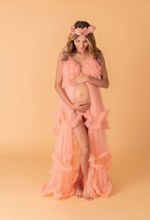 Maternity photo of woman in pink ruffled dress with floral crown against peach backdrop.