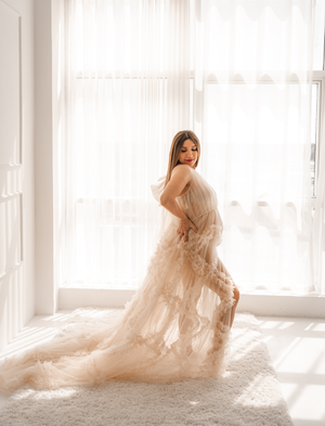 Woman in flowing beige dress poses by sunlit window in bright white room.