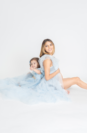 Mother and daughter in matching blue dresses sit back-to-back on white backdrop.