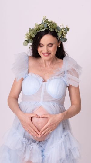 Pregnant woman in light blue tulle dress wears eucalyptus crown while making heart shape on belly.