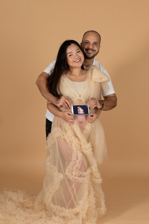 Expectant couple holds ultrasound photo while embracing in front of brown backdrop wearing cream-colored outfits.