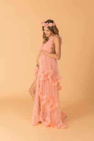 Elegant maternity session in flowing coral dress against warm backdrop.