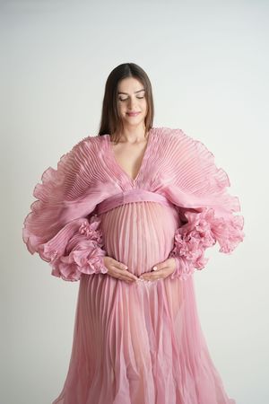Woman in flowing pink pleated maternity gown with dramatic ruffled sleeves poses against white backdrop.