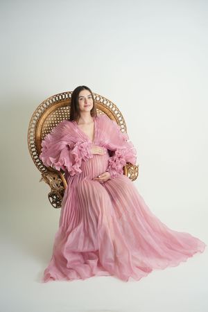 A dreamy maternity portrait in a blush pink pleated gown with dramatic ruffled sleeves on an ornate cane chair.