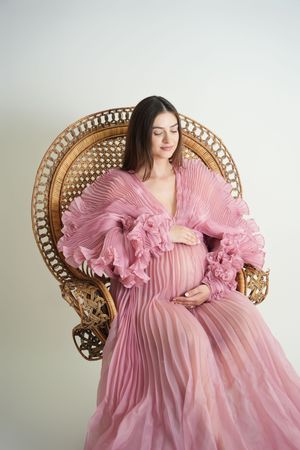 A romantic maternity photo featuring a pink pleated gown with dramatic ruffled sleeves on a vintage wicker chair.