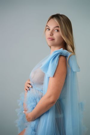 Expectant figure wearing romantic light blue tulle gown with bow detail against neutral backdrop.