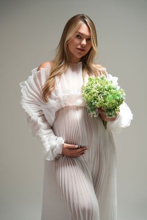 Figure in a flowing white pleated dress poses elegantly with small green floral bouquet against neutral backdrop.
