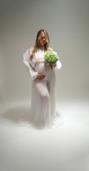 A figure in a flowing white pleated dress stands against a light background holding a bouquet of fresh flowers.