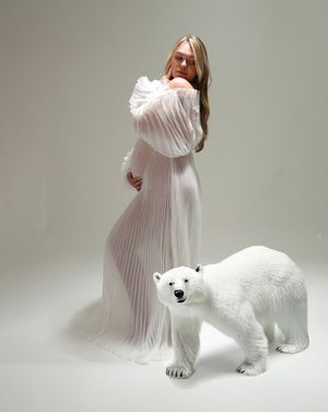 A person in a white pleated dress poses with a large white polar bear statue against a light gray backdrop.