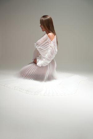 Figure in ethereal white pleated dress poses gracefully while sitting in soft studio lighting.