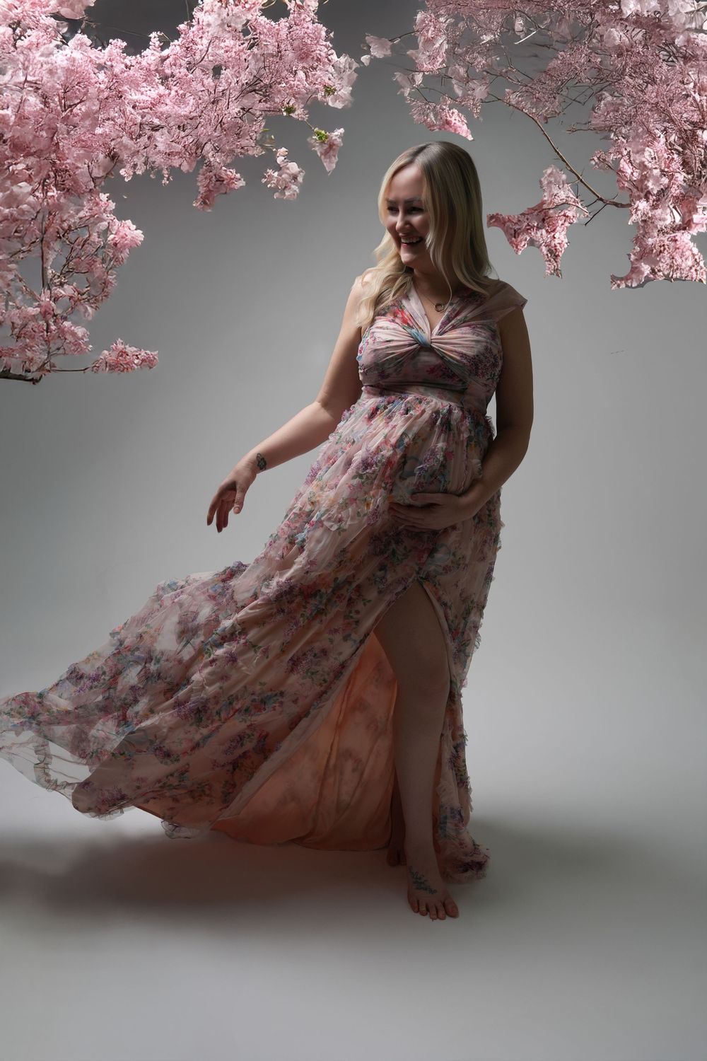 A floral dress flows gracefully under pink cherry blossoms against a gray studio backdrop in an artistic portrait series.