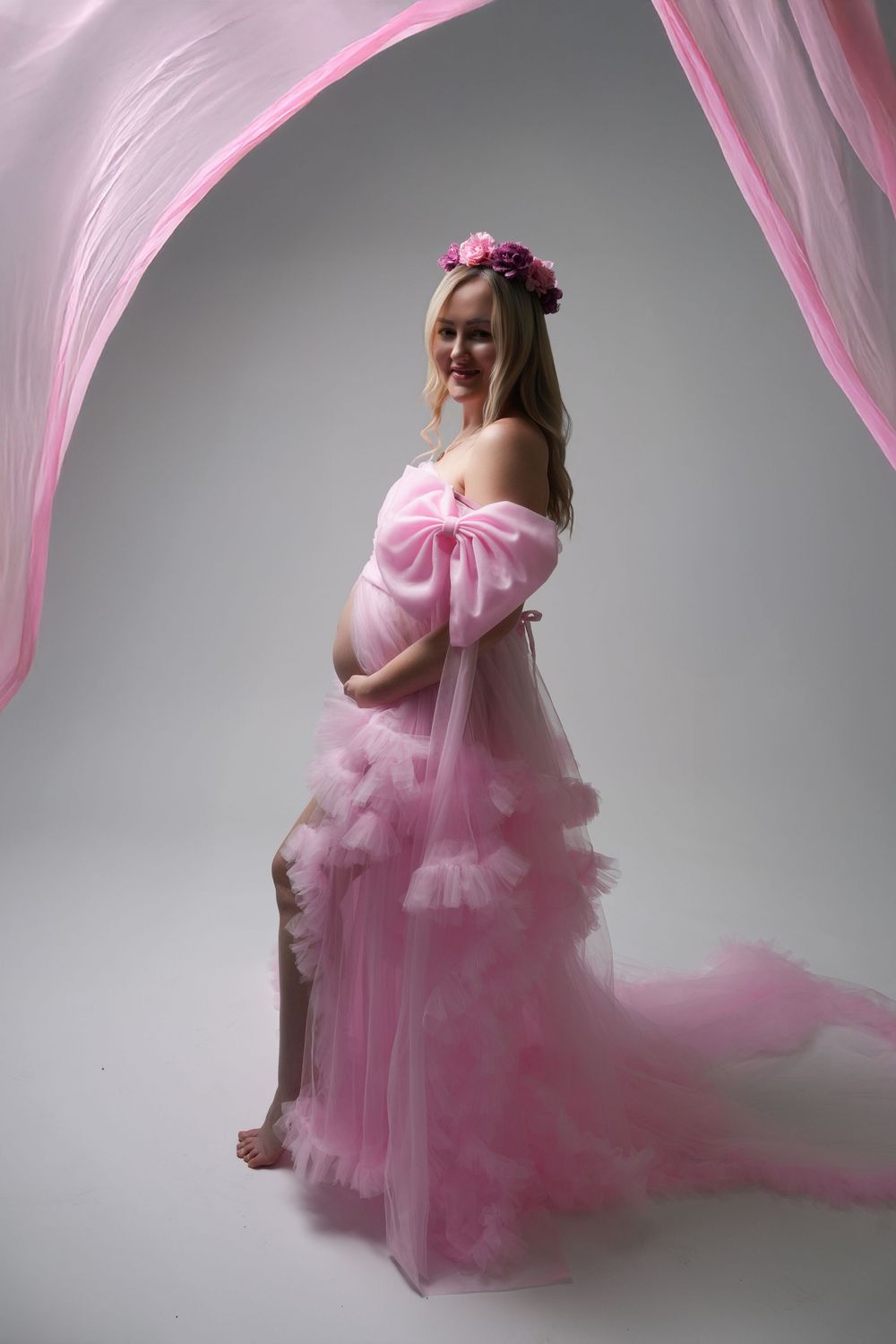 Maternity portrait with flowing pink tulle dress and floral crown against gray background in dreamy artistic style.