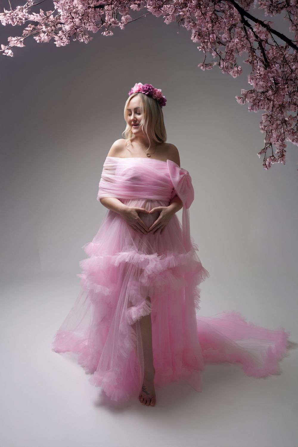 A pink tulle gown with off-shoulder design photographed under cherry blossoms against a gray backdrop.