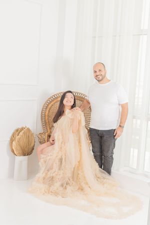A couple relaxes in a wicker chair with flowing peach-colored fabric in an airy white room.