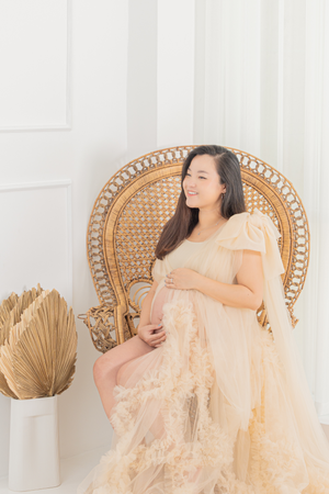 Pregnant woman in cream lace dress sits on rattan peacock chair with dried palm decor.