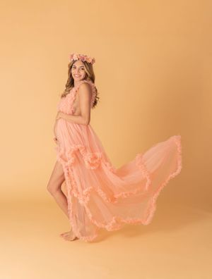 Graceful maternity poses in flowing coral dress against peachy backdrop.