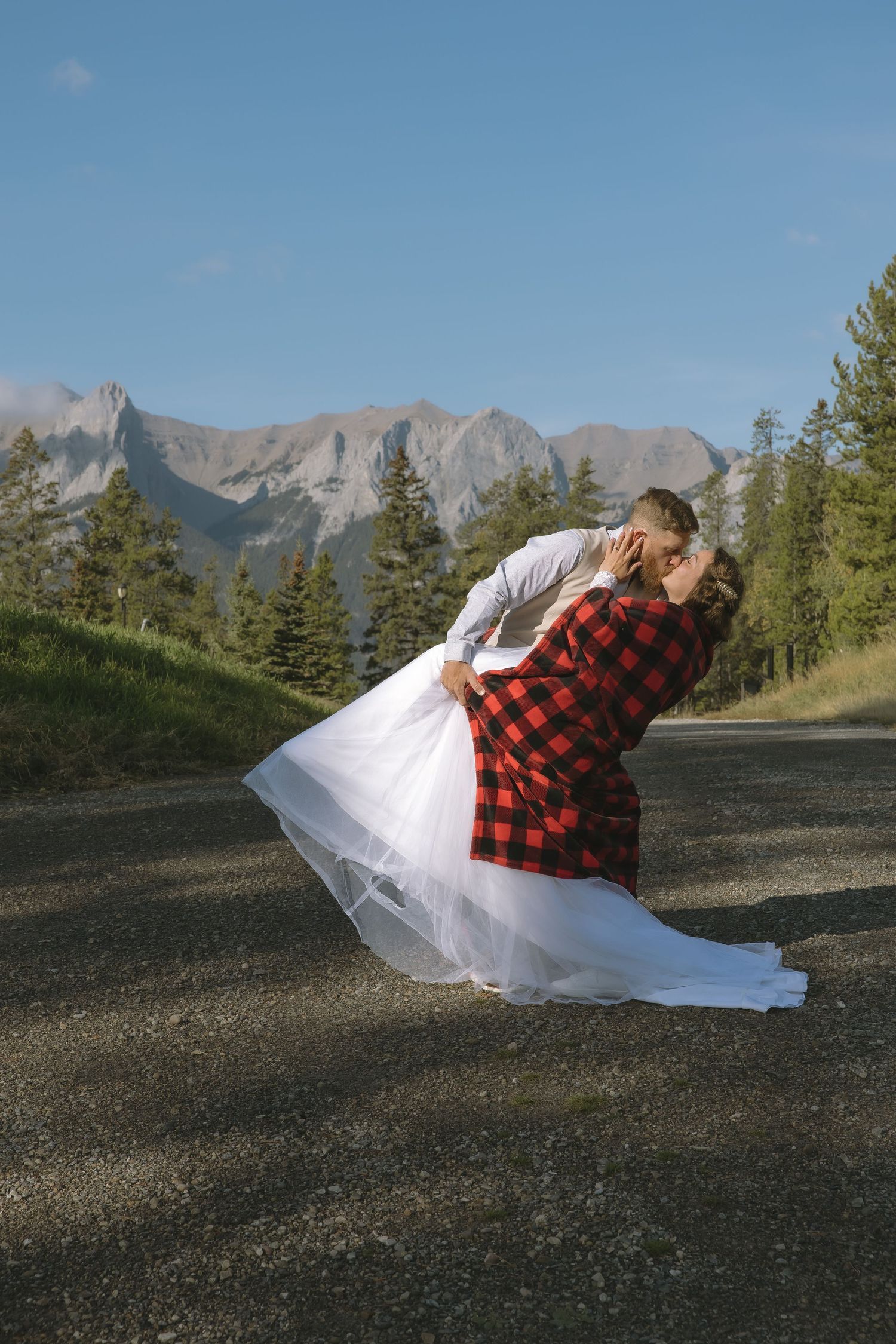 A couple in wedding attire and red plaid flannel embrace against a scenic mountain backdrop in a creative photo series. Minot, ND photograph