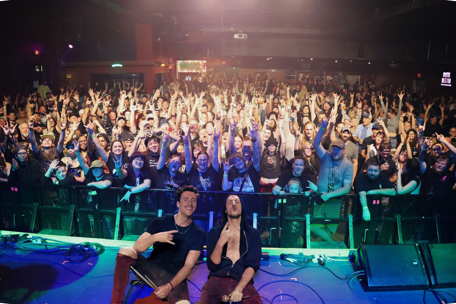 A large concert crowd with raised hands and colorful stage lighting while performers sit at the edge of the stage.