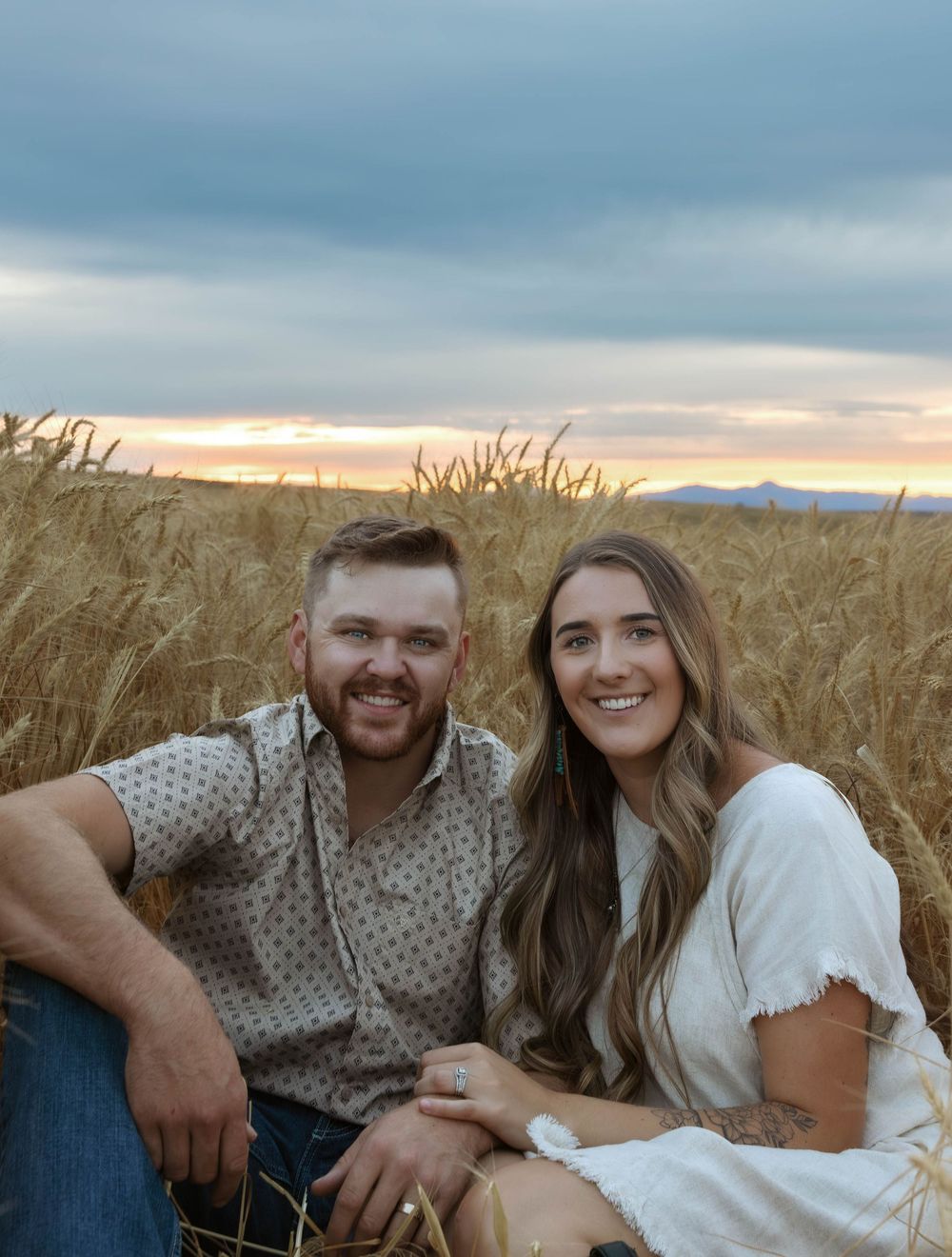 A romantic photoshoot in a wheat field with cloudy skies and sunset in the background.