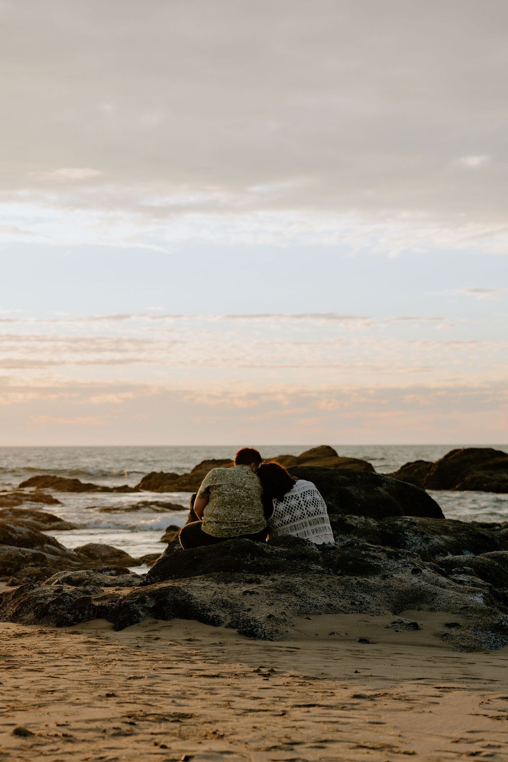 A couple sits on a rocky beach at sunset with waves crashing against the shore.
