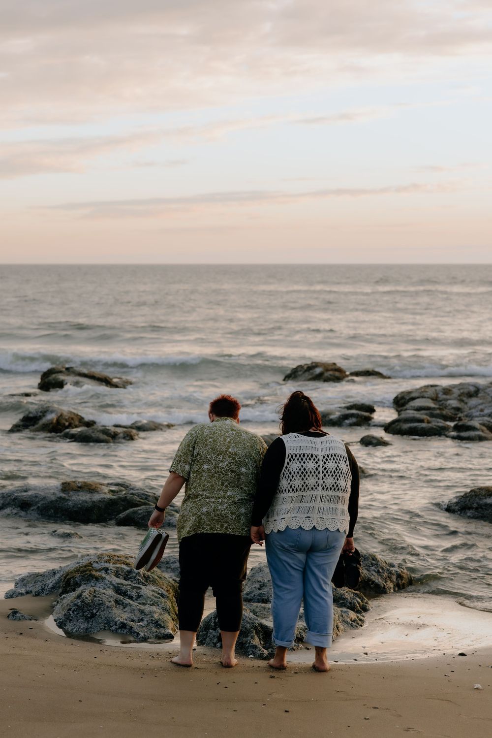 Two people stand together watching the ocean waves from a rocky beach at sunset.